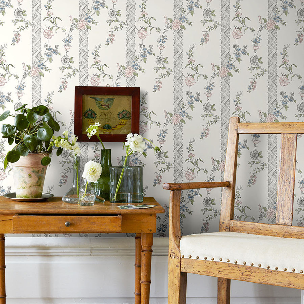 Vintage wooden table with drawer, glass vases and potted plant beside a weathered chair; floral wallpaper backdrop.