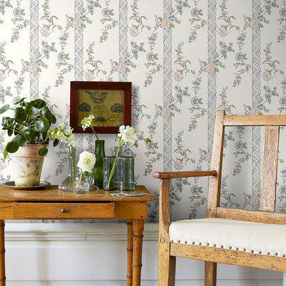 Vintage wooden table with drawer, glass vases and potted plant beside a weathered chair; floral wallpaper backdrop.