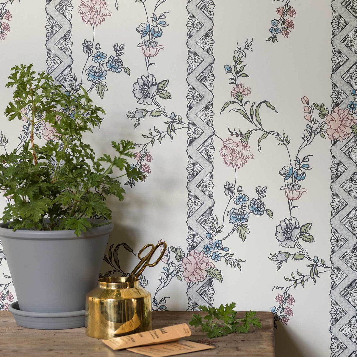 Green leafy plant in a gray pot on a wooden table, with a small gold container beside it, floral wallpaper backdrop.