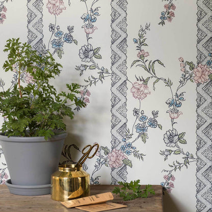 Green leafy plant in a gray pot on a wooden table, with a small gold container beside it, floral wallpaper backdrop.