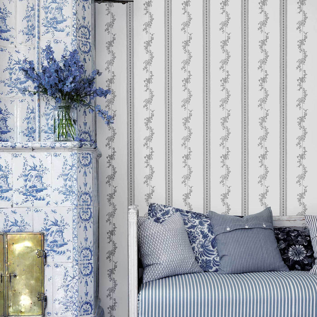 Blue and white chinoiserie scene with tiled fireplace, glass vase of blue flowers, striped bedding and patterned cushions.