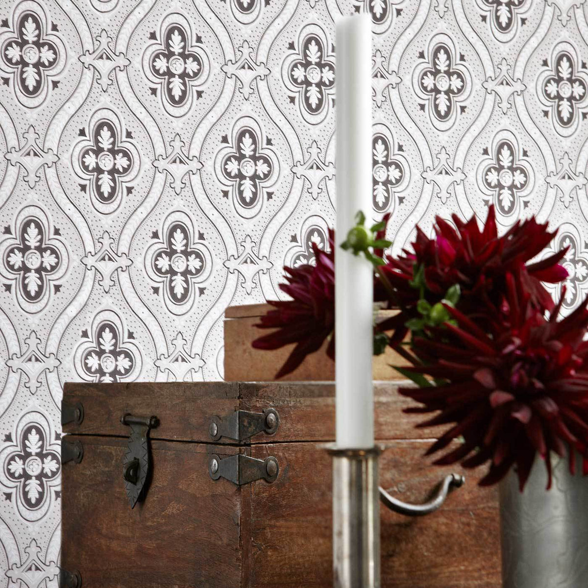 Tall white candle in a silver holder beside a burgundy bouquet on a rustic wooden chest against damask wallpaper.