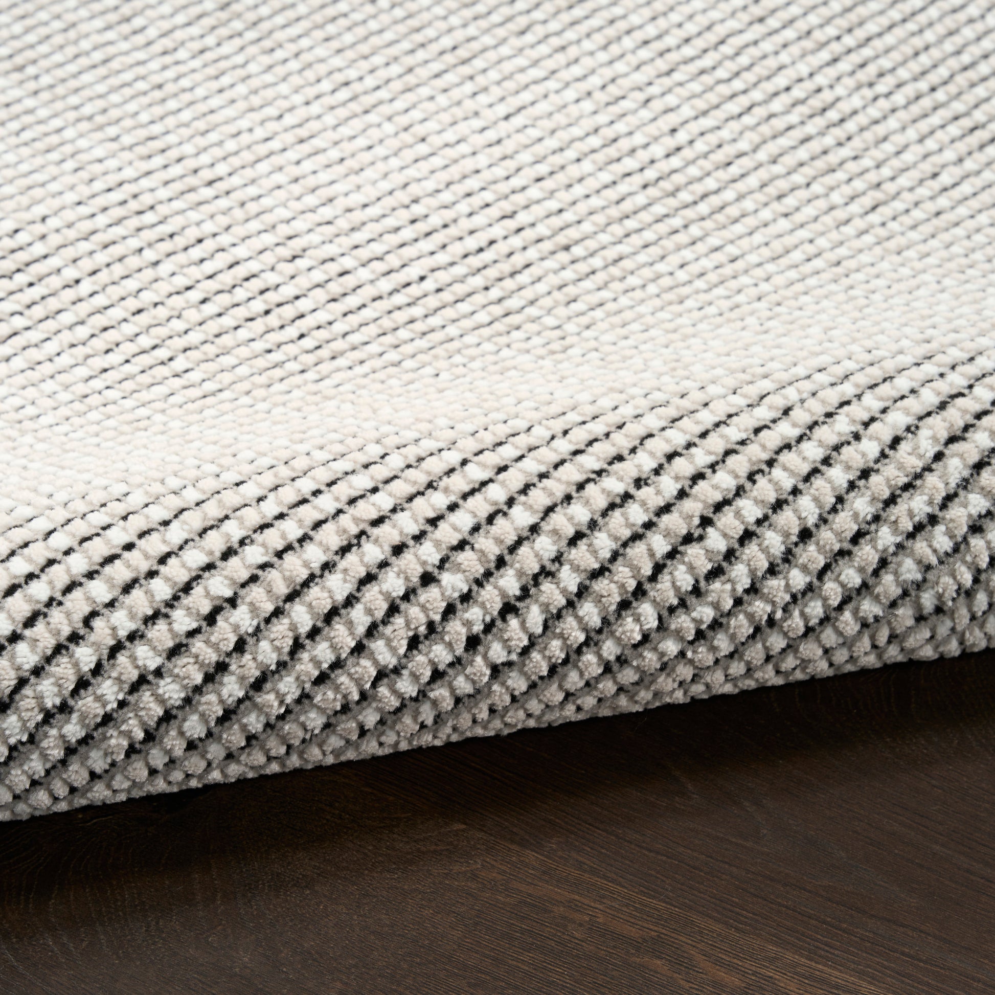 Beige textured woven rug with subtle black checkered pattern, edge curled on dark wood floor.