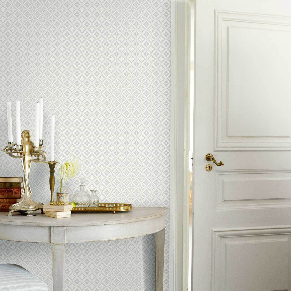 Round marble-top console with gold trim, candlestick, books, vase of white flowers; diamond wallpaper and white paneled door.