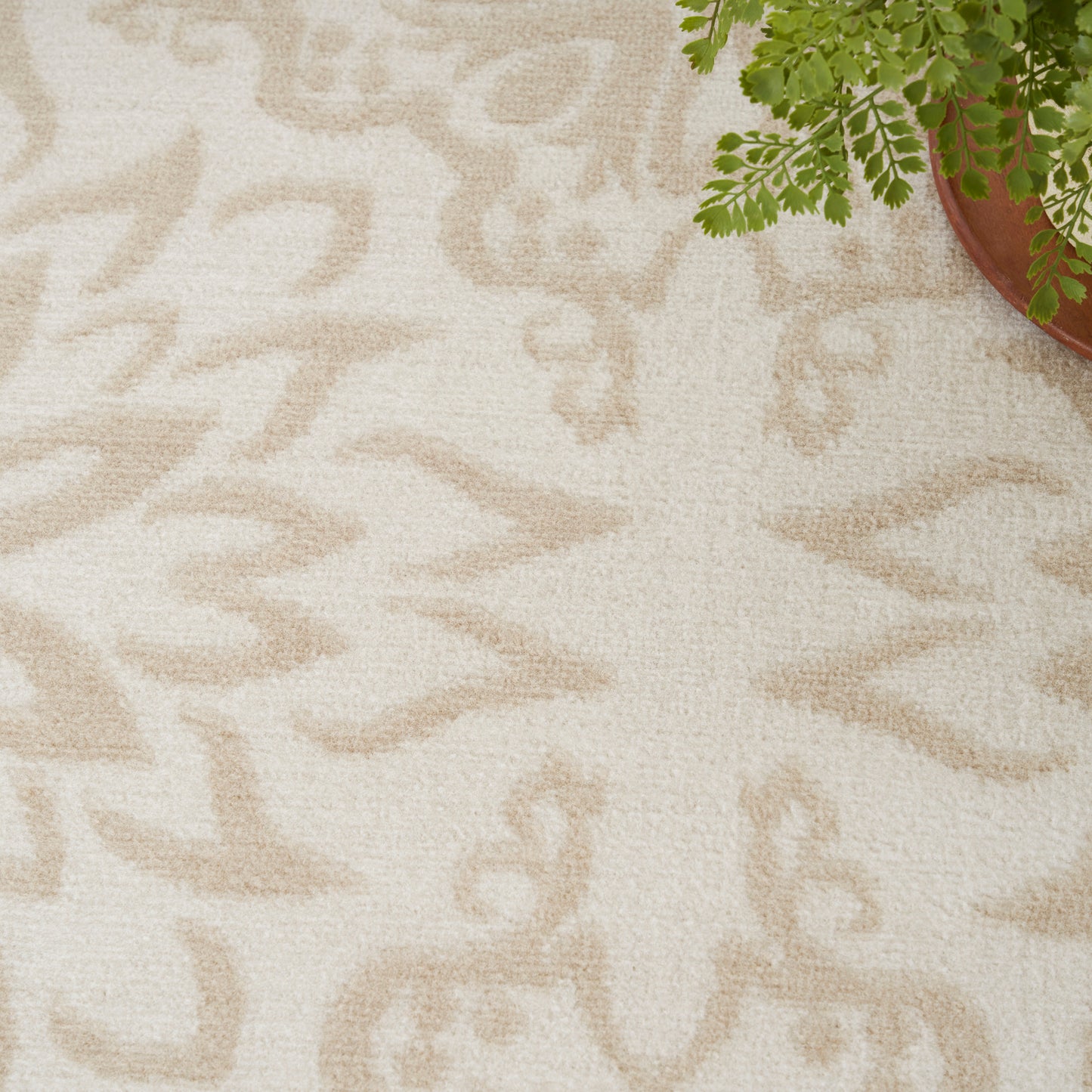 Cream rug with a light tan abstract leaf pattern and textured weave; partial view of a potted fern in the corner.