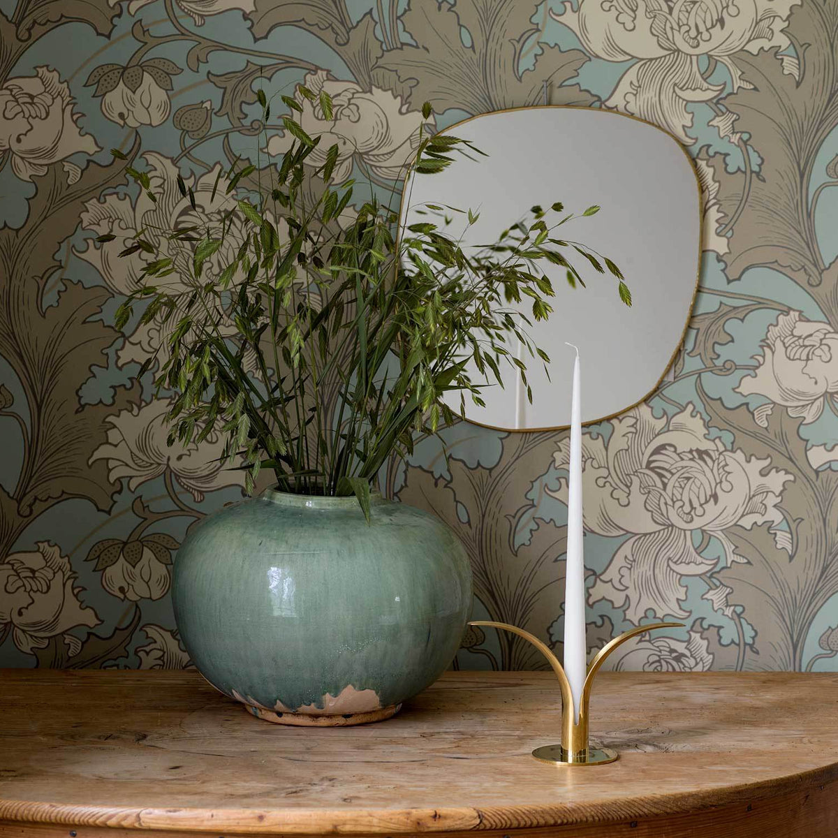Teal ceramic pot with bamboo on a wooden table, white taper candle in brass holder, circular mirror and wallpaper.