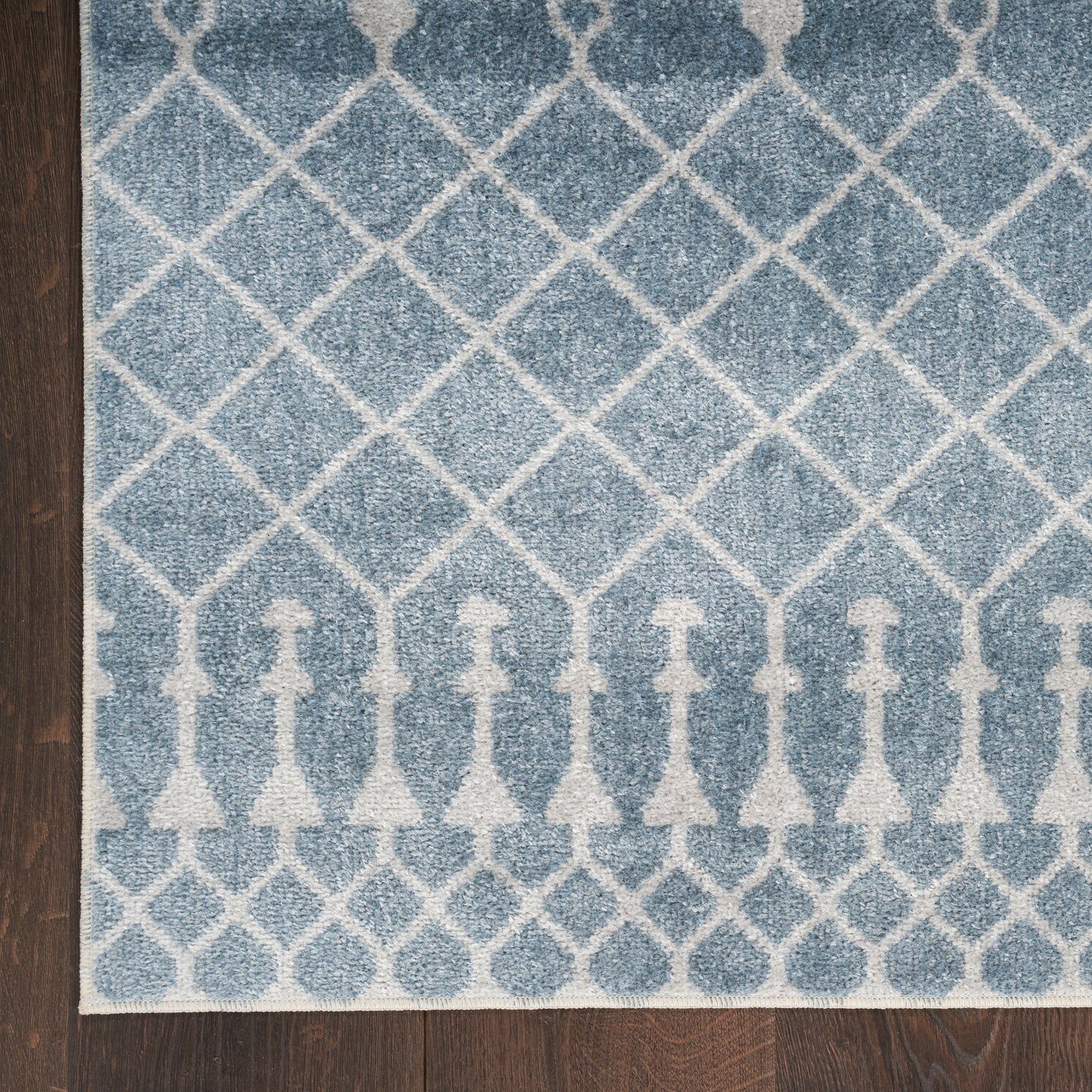 Blue woven rug with white lattice diamond pattern and decorative border motifs, on dark wooden floor.