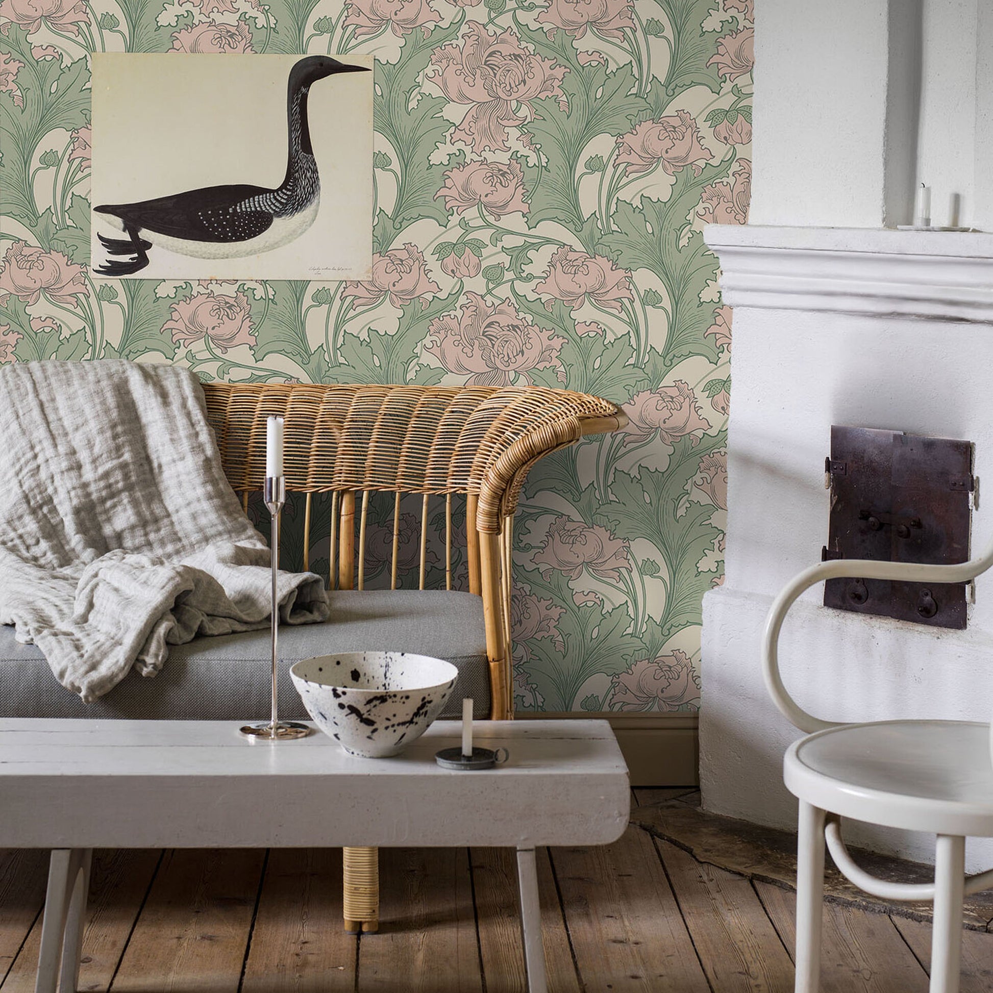 Rustic living room with wicker armchair, gray sofa, white coffee table, botanical green-pink wallpaper, candles, and ceramic bowl.