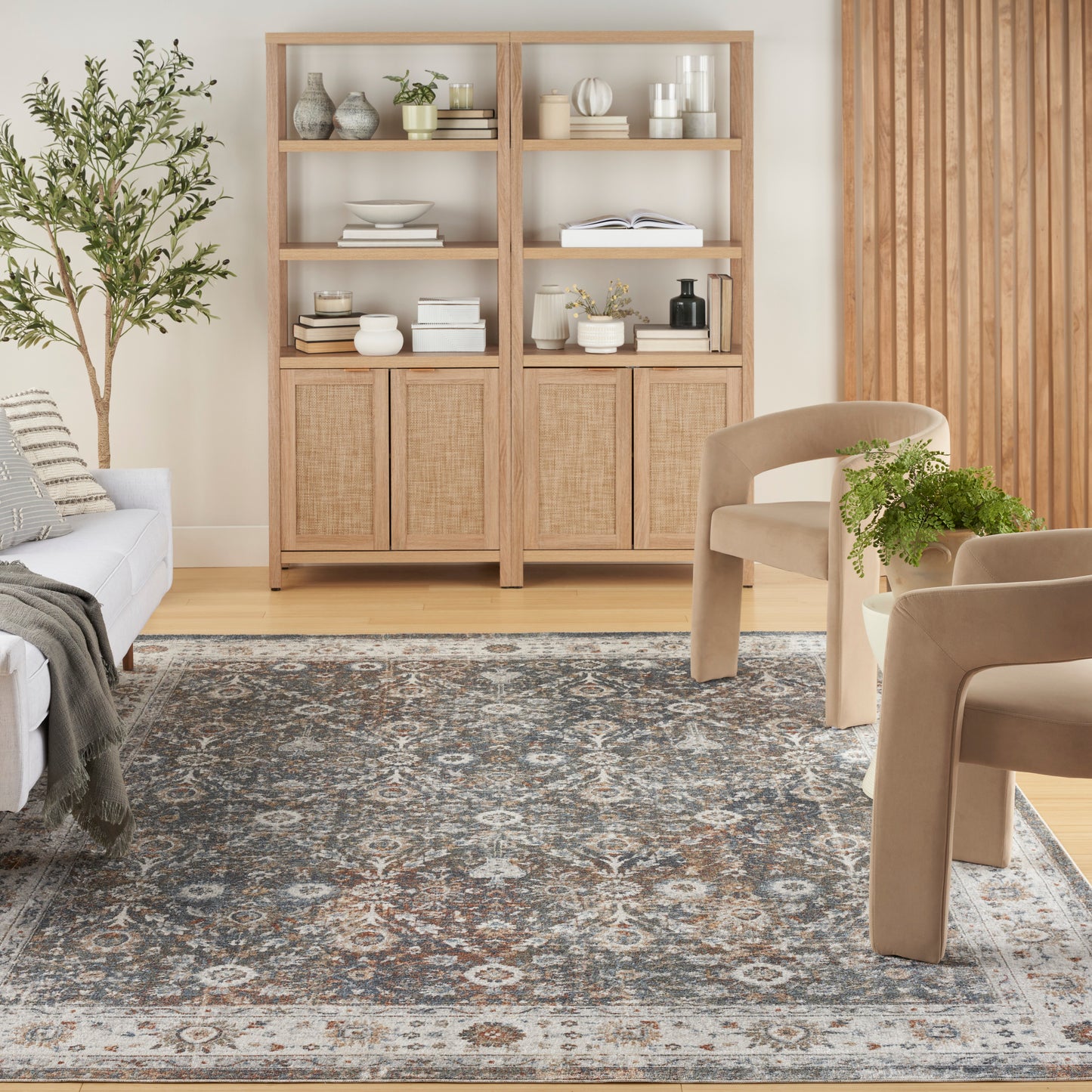 Light oak shelving unit with woven rattan doors, open shelves with vases and books; beige lounge chairs and a blue-gray patterned rug.