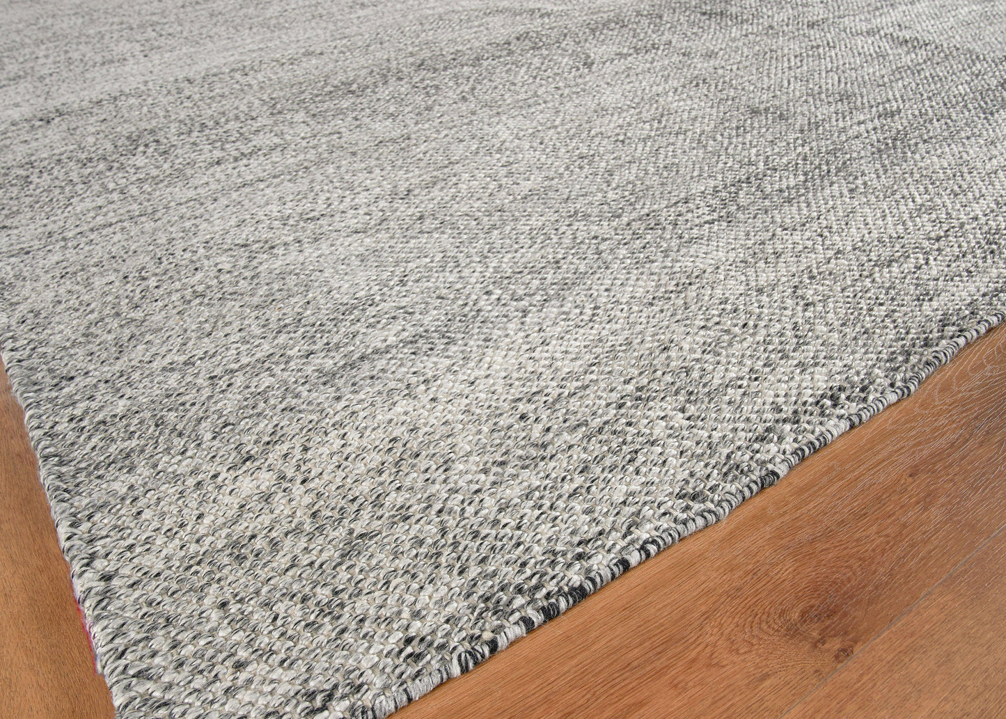 Grey speckled woven rug with dark binding edge, low-pile modern rectangular area rug on wood floor.