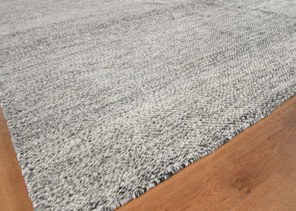 Grey speckled woven rug with dark binding edge, low-pile modern rectangular area rug on wood floor.
