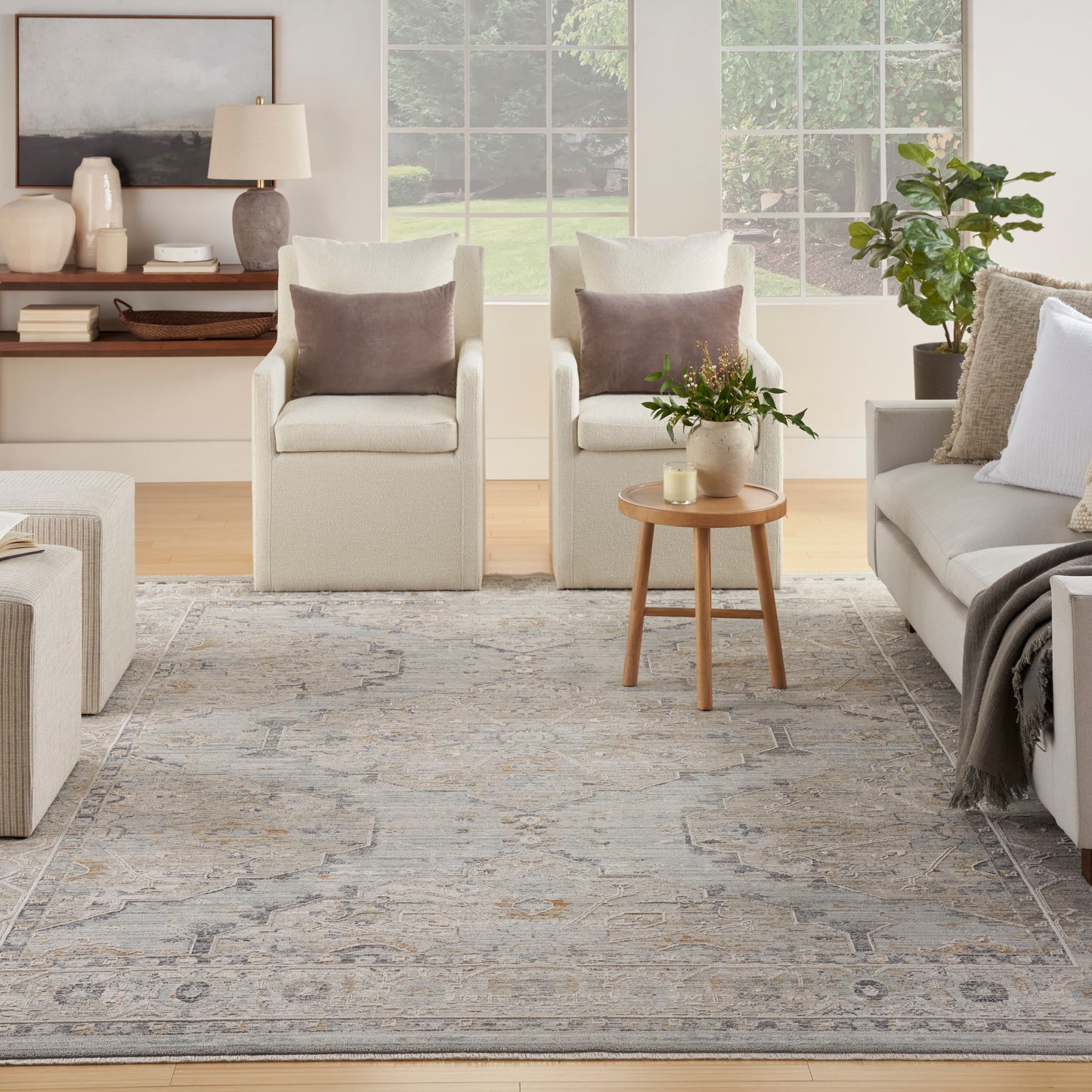 Neutral living room with cream armchairs, brown pillows, beige sofa, round wood side table with plant and candle, gray rug.