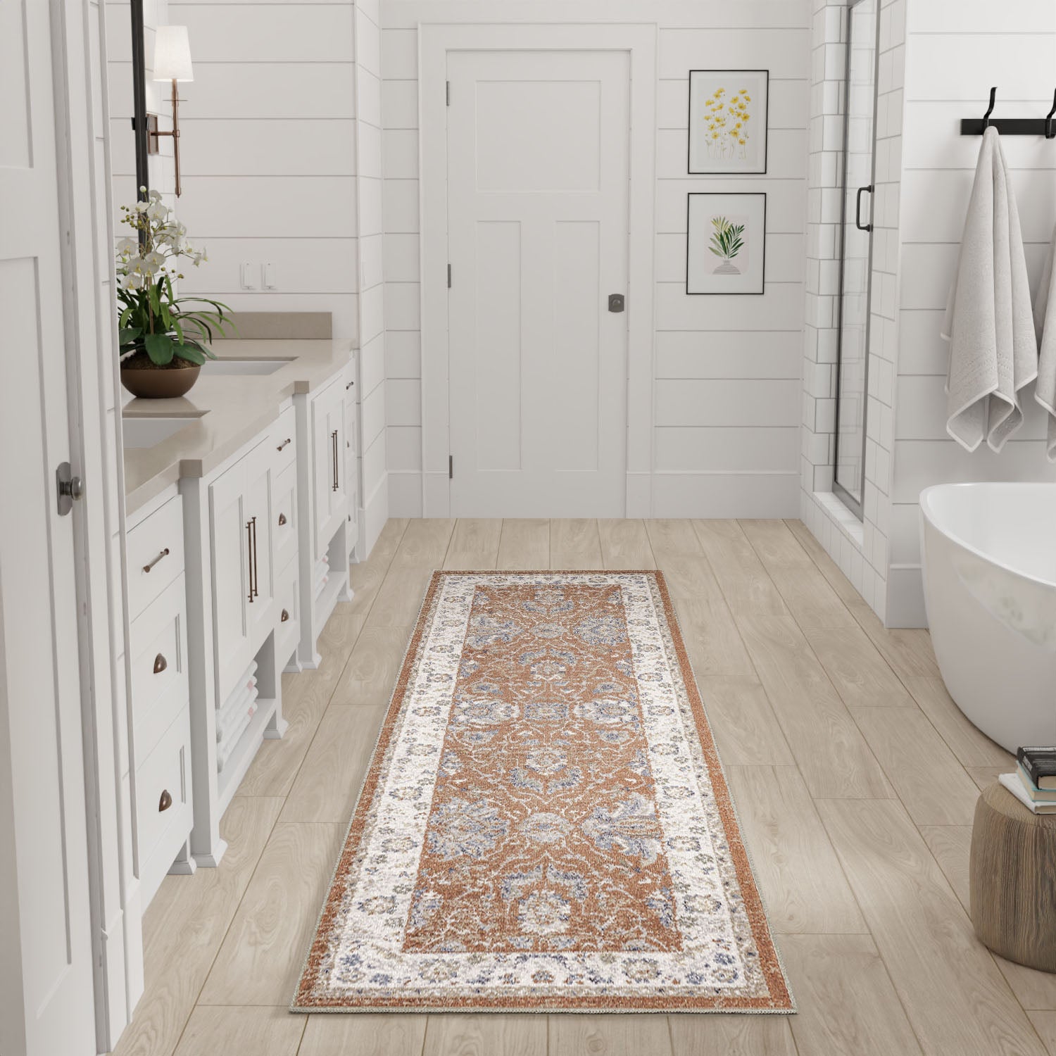 Long orange-and-blue oriental runner rug in a bright white bathroom with light wood floors, white cabinetry, plant, and tub.