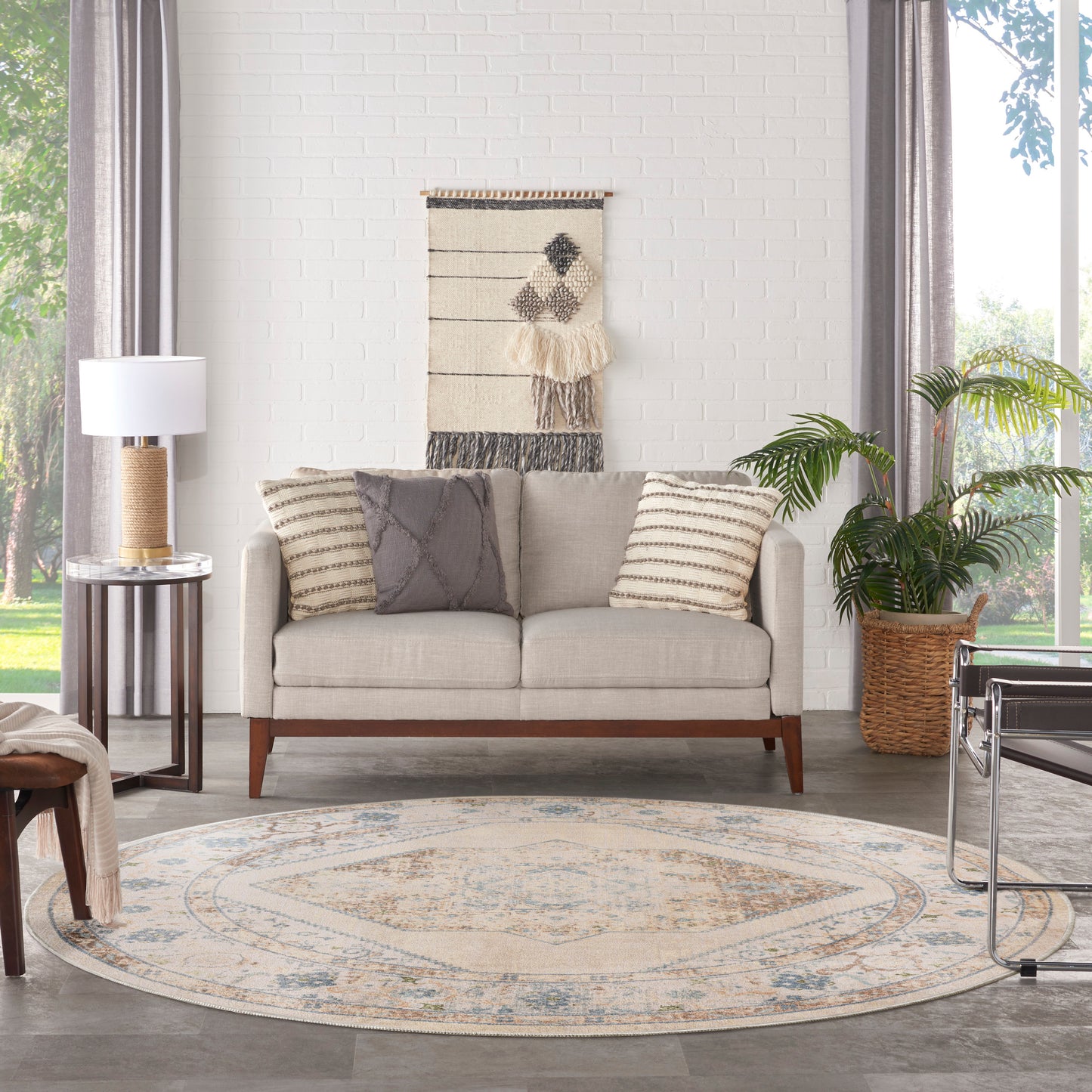 Beige sofa with striped pillows, round patterned rug, woven wall hanging, plant, and glass side table by large window.