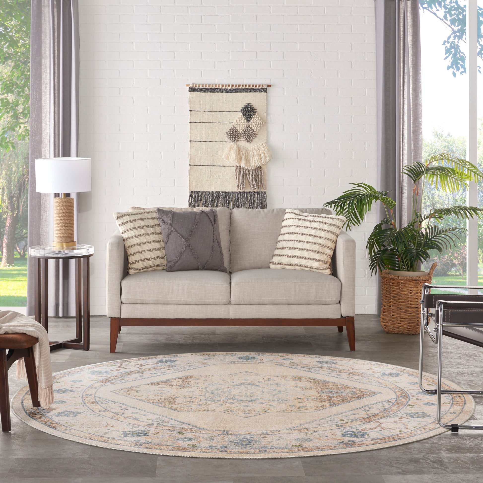 Beige sofa with striped pillows, round patterned rug, woven wall hanging, plant, and glass side table by large window.
