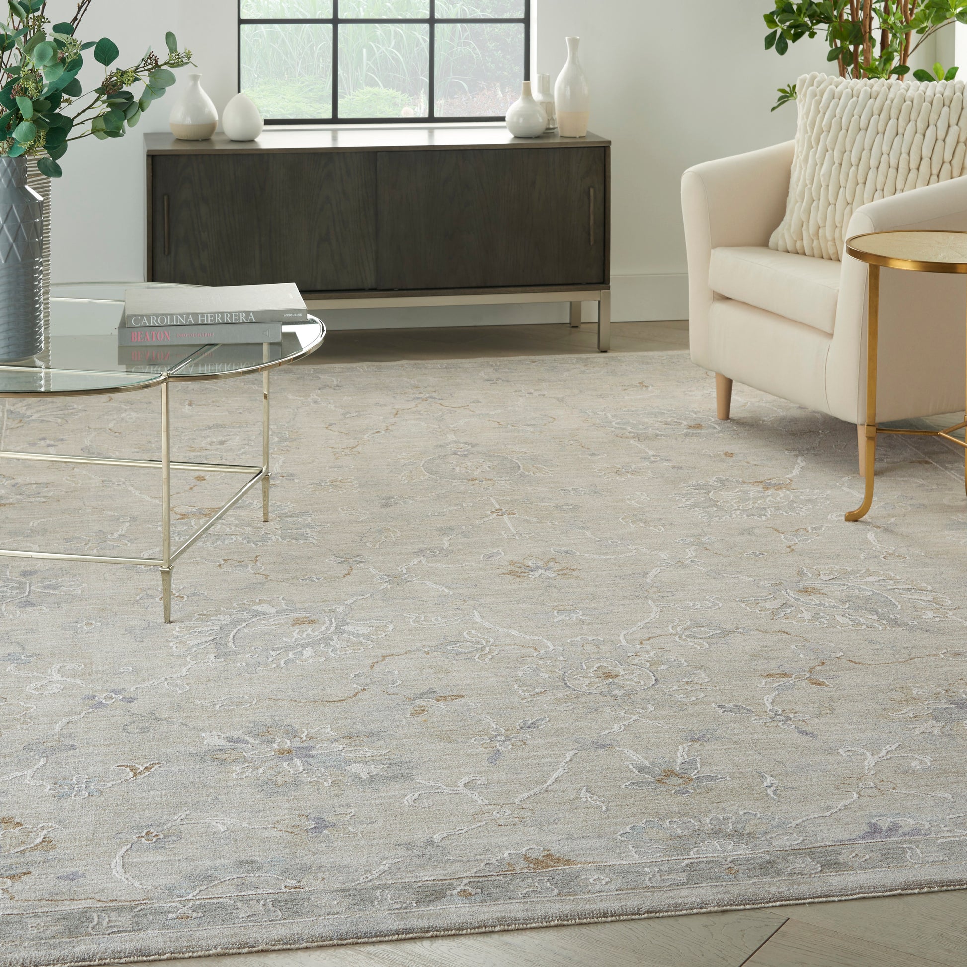 Soft gray ornate area rug with beige floral motif; glass coffee table, cream sofa with gold legs, dark wood sideboard.