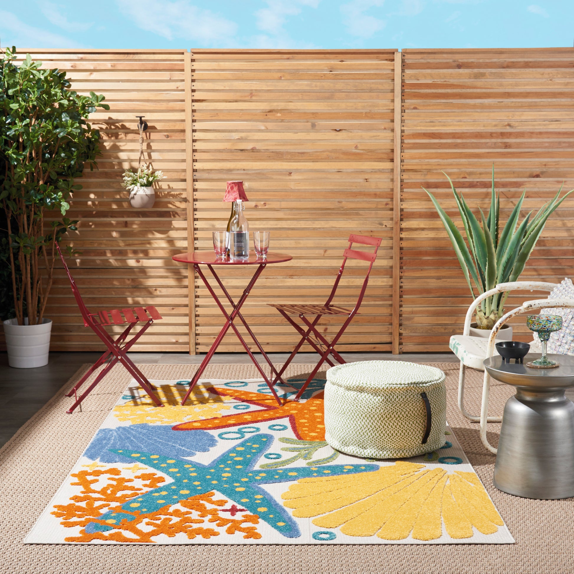 Colorful starfish-shell coastal rug on a porch, with a red metal bistro set, a woven pouf, and potted plants against a wooden fence.