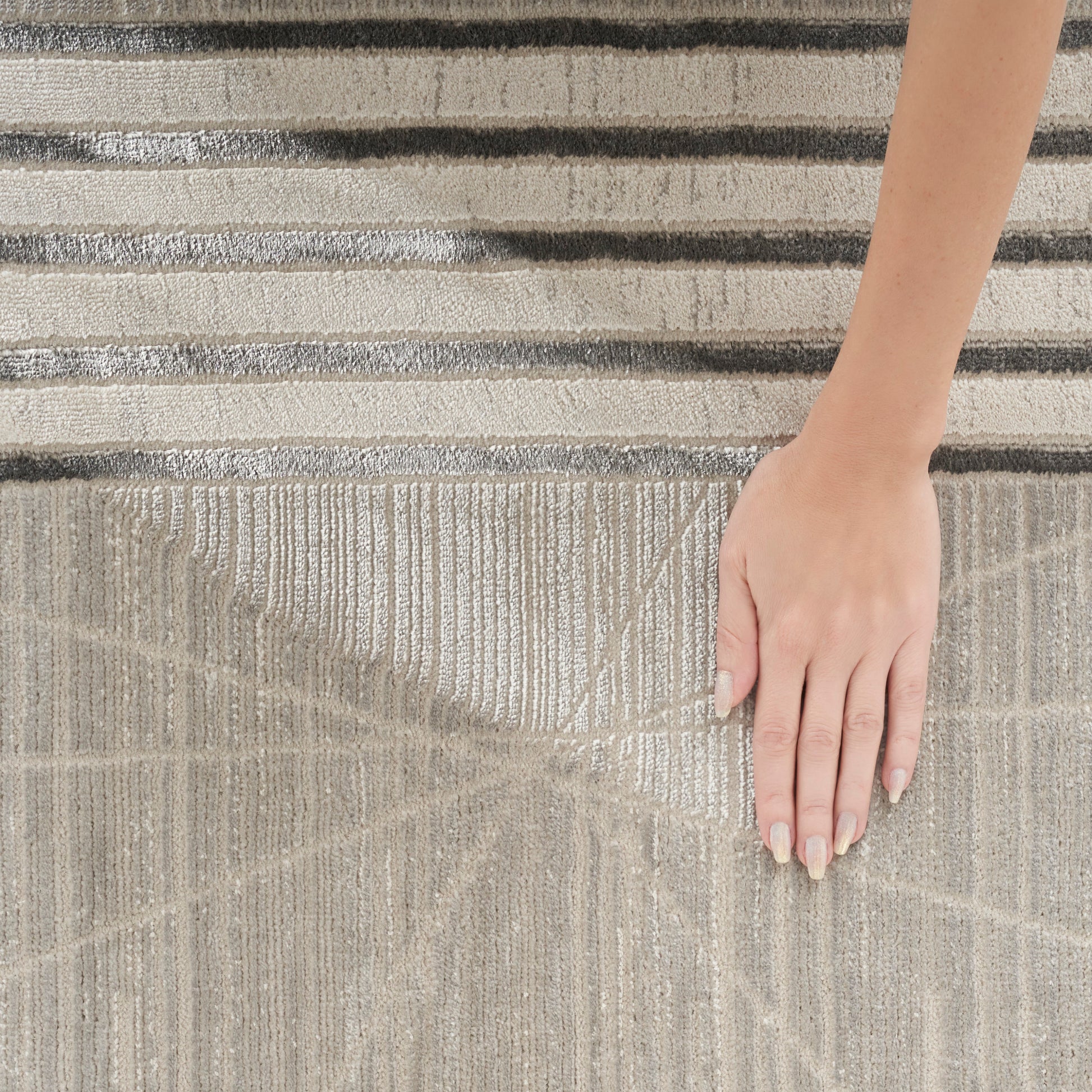 Beige textured area rug with geometric line patterns; a hand with neutral manicured nails rests on it, striped rug in background.