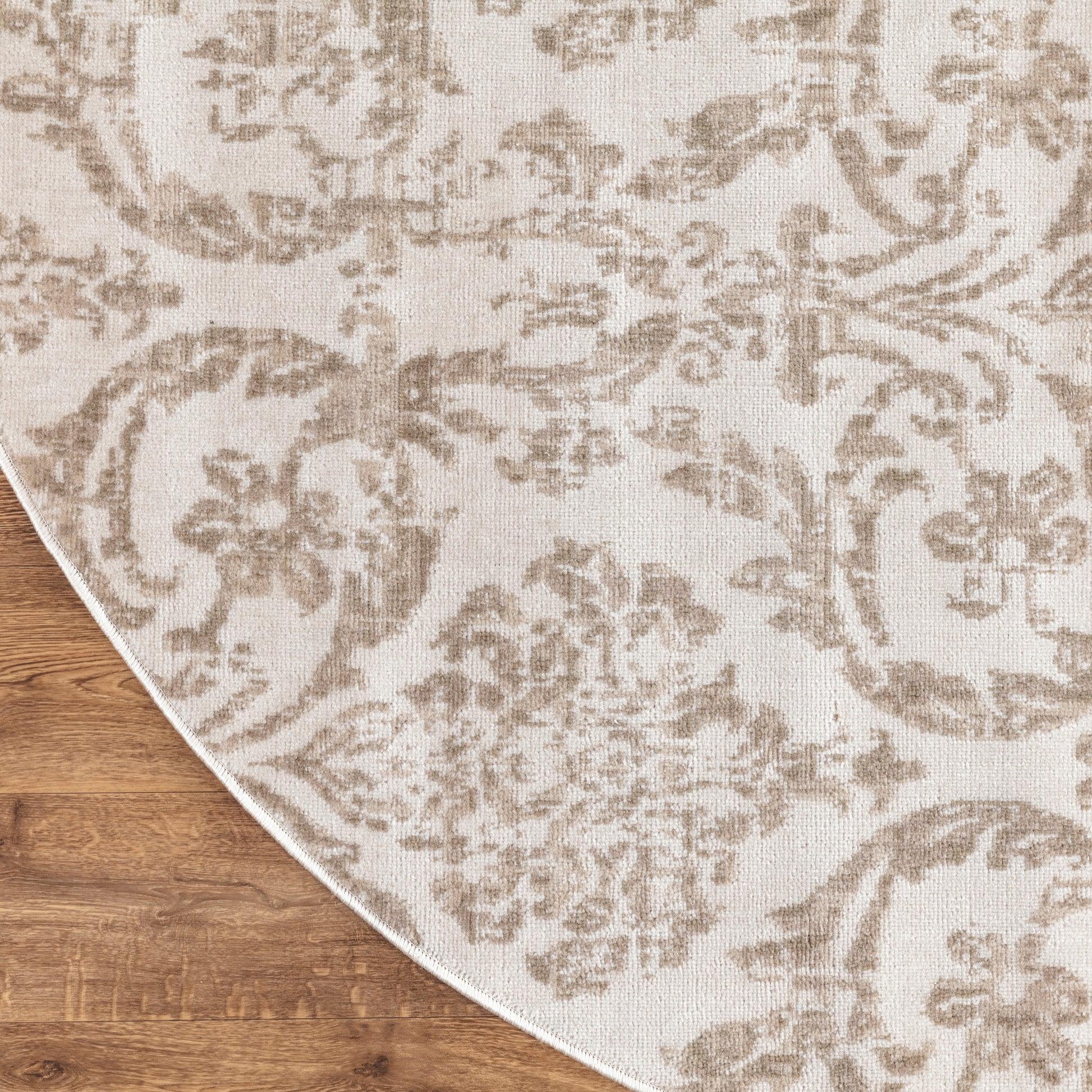 Beige damask-patterned round area rug with taupe floral motif on wood floor; curved edge visible.