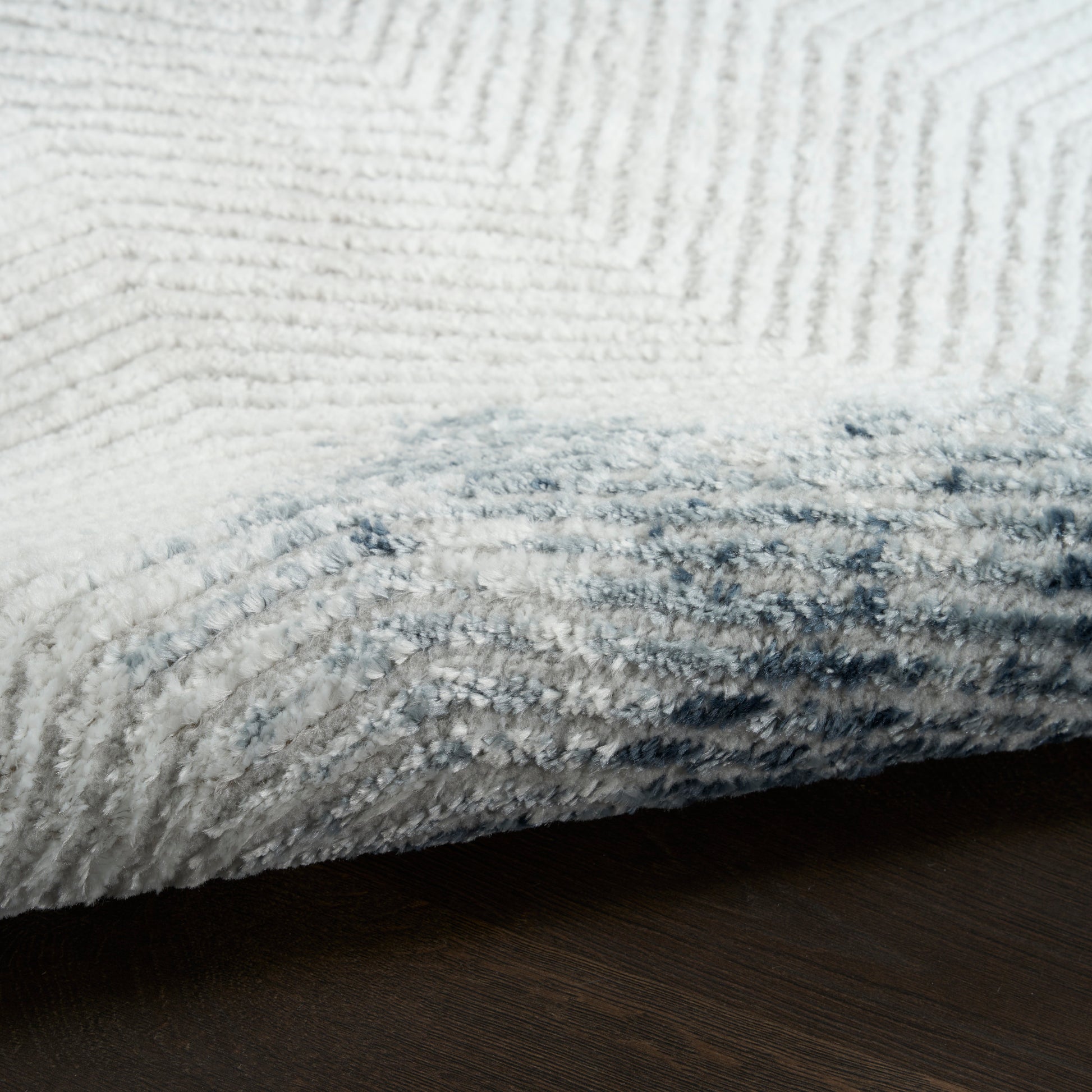 White plush rug with thick pile and light gray-blue edge, rolled corner on dark wood floor.