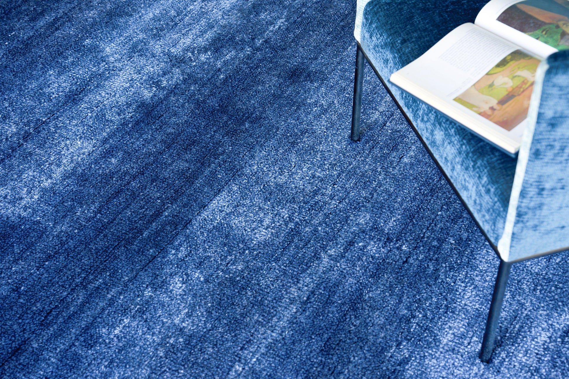 Blue textured rug with a blue velvet chair & slim black legs; open magazine resting on chair seat.