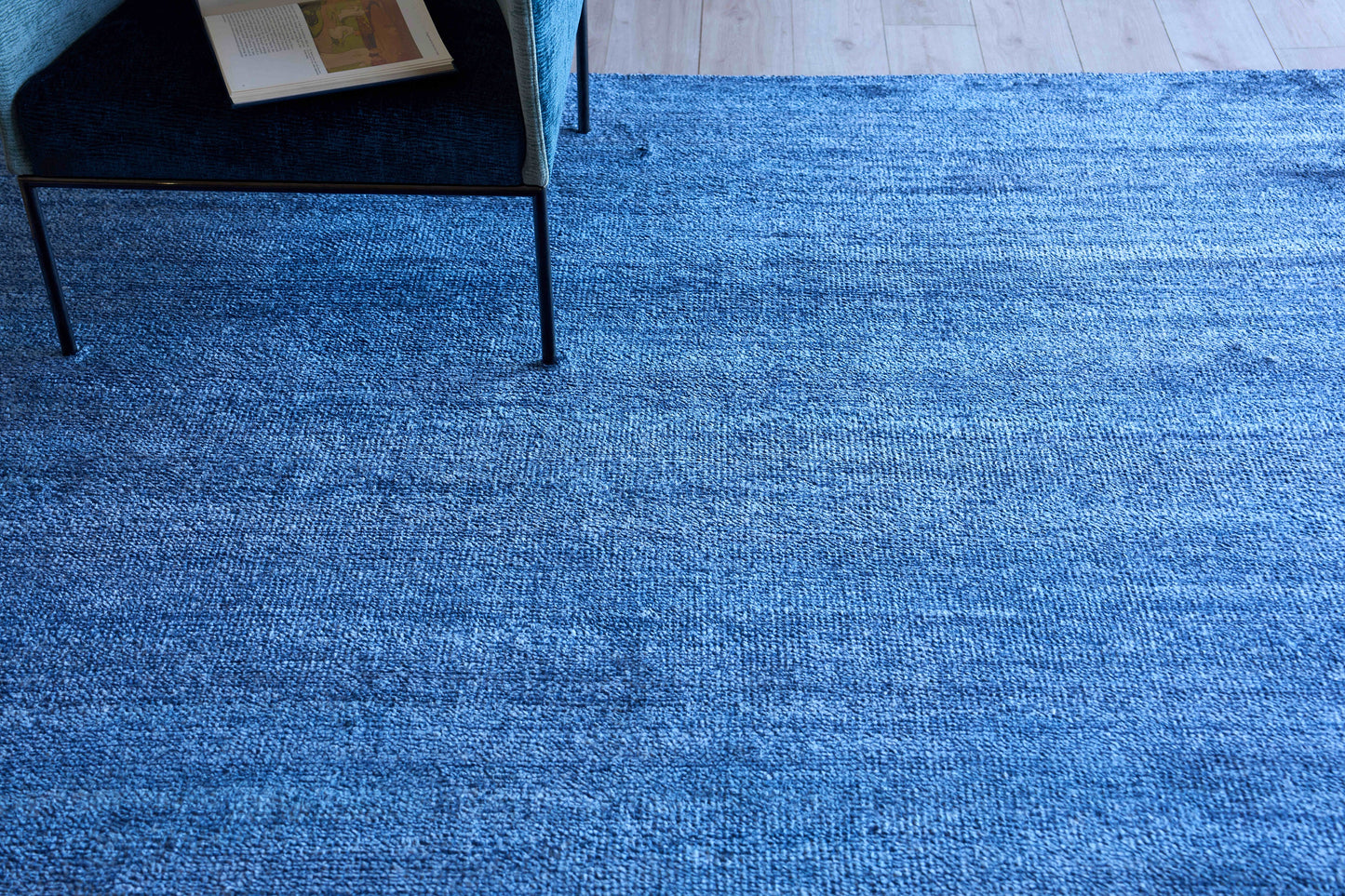 Blue textured fabric rug on light wood floor; modern chair with black metal legs at edge, magazine on seat.