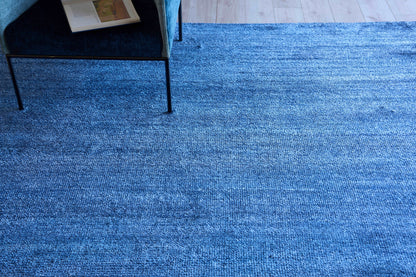 Blue textured fabric rug on light wood floor; modern chair with black metal legs at edge, magazine on seat.