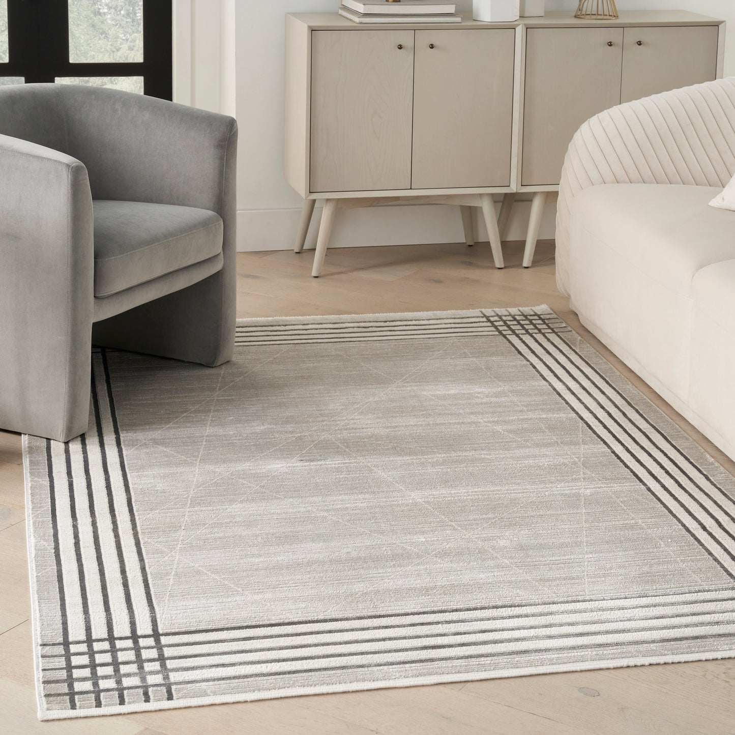 Light gray area rug with dark striped border in a modern living room with a gray armchair and beige sofa.