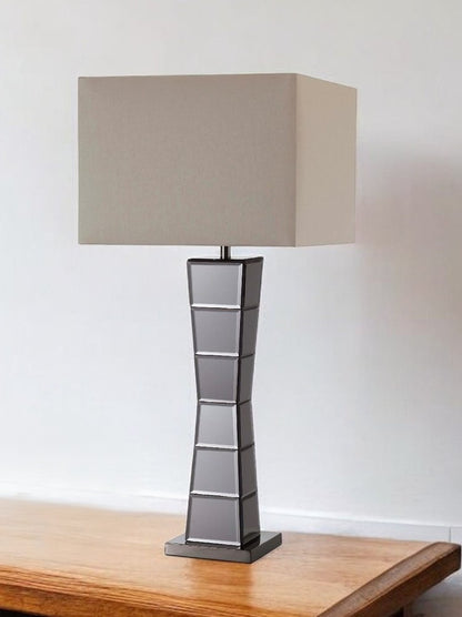 Black glass tower table lamp with a beige fabric shade on a table.