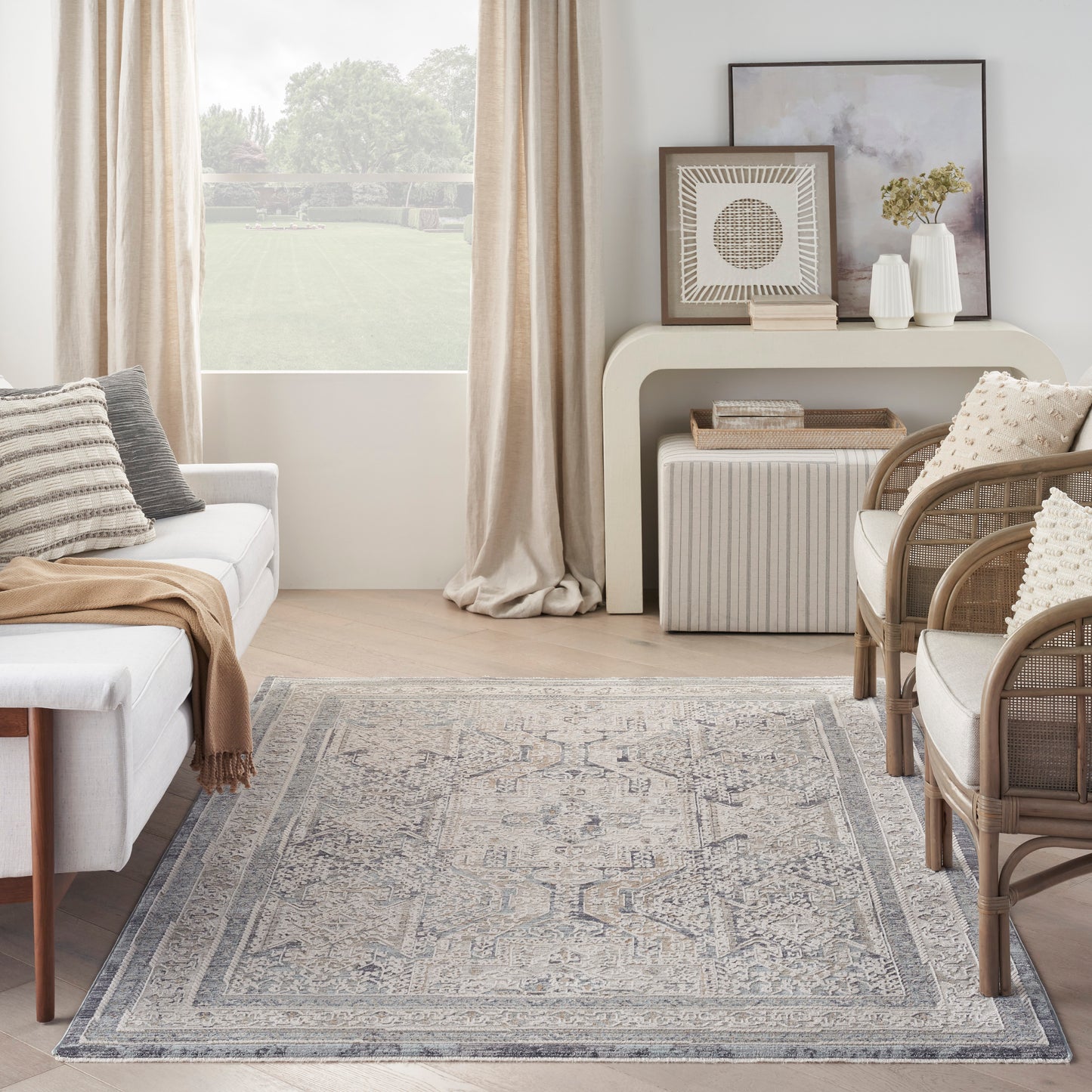 Neutral living room with blue-gray rug, white sofa with beige throw, wicker chairs, beige curtains, and a console with art.