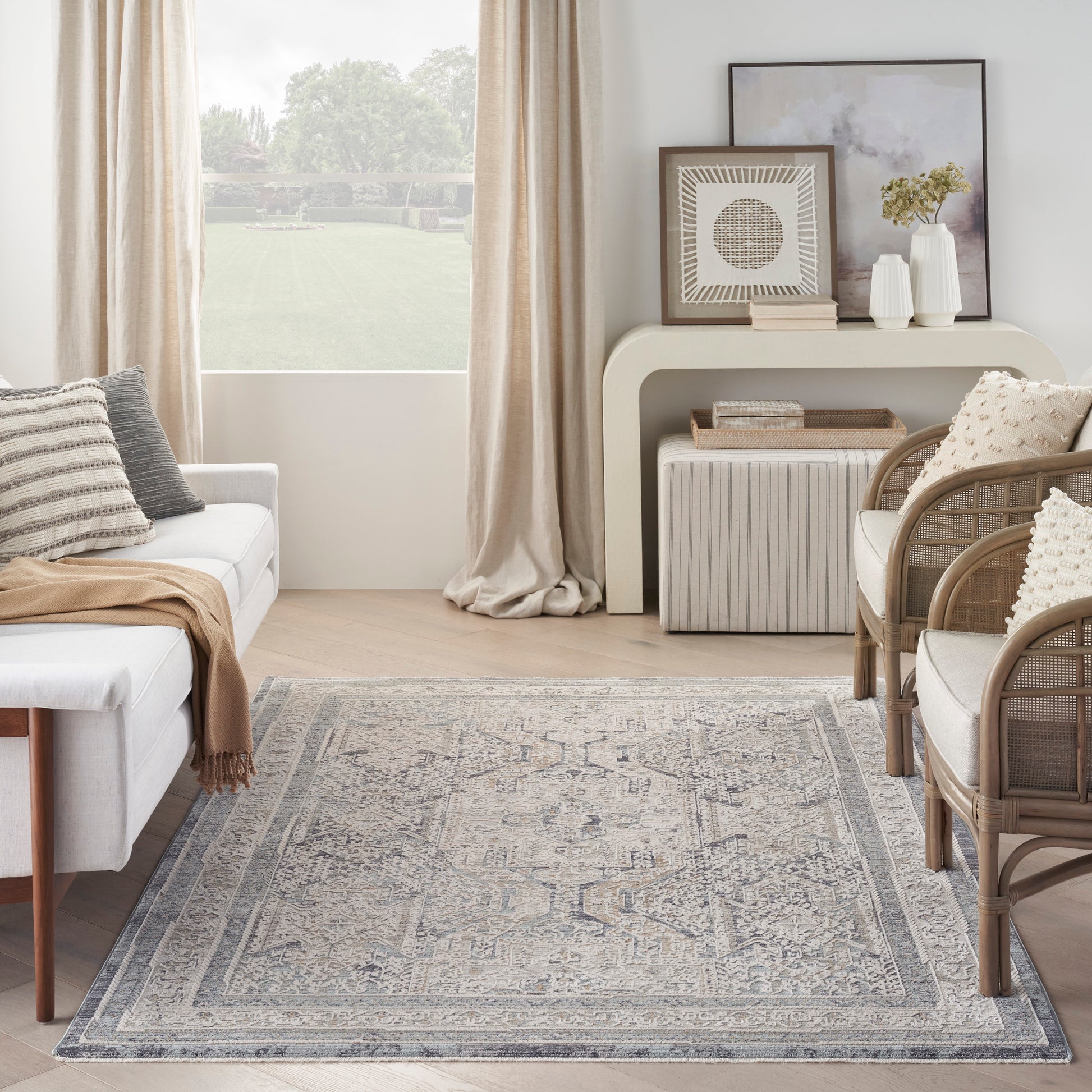Neutral living room with blue-gray rug, white sofa with beige throw, wicker chairs, beige curtains, and a console with art.