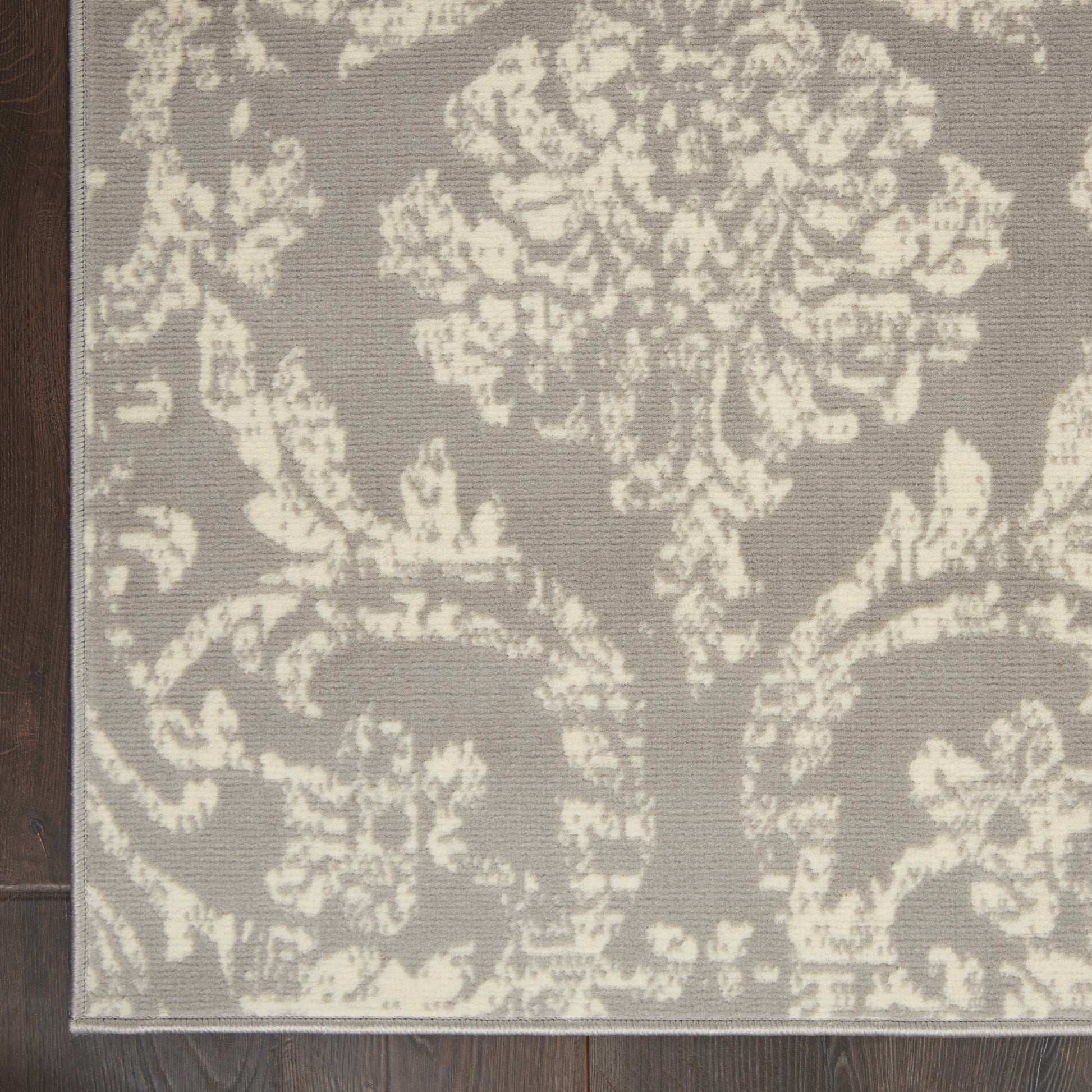 Taupe rug with cream damask floral pattern on dark wood floor.