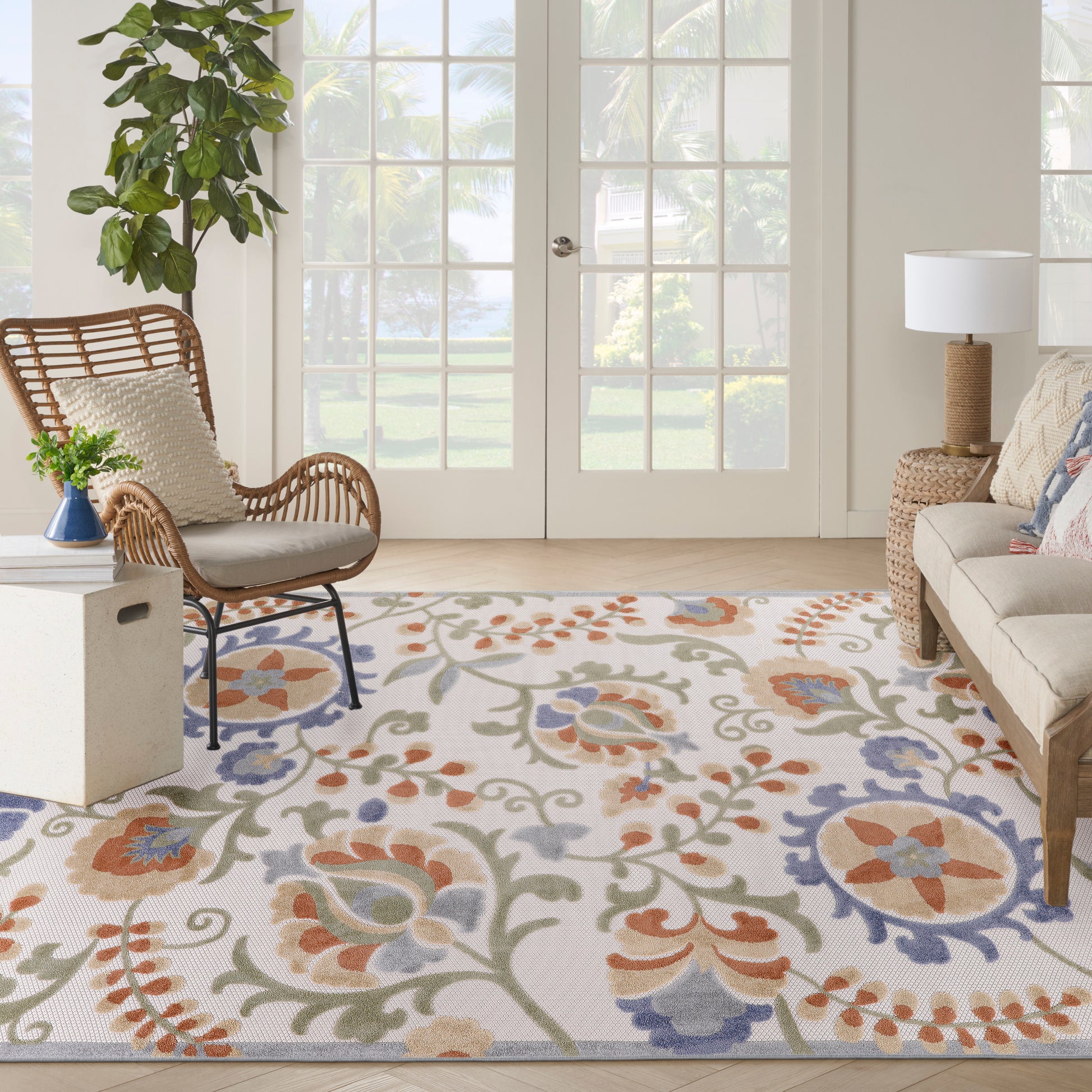 Bright living room with a colorful floral area rug, a rattan chair, beige sofa, potted plant, and glass French doors.