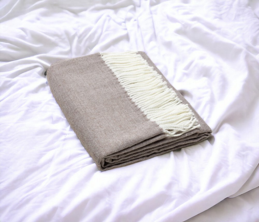 Taupe knit throw blanket with white fringe, neatly folded on a white bed.