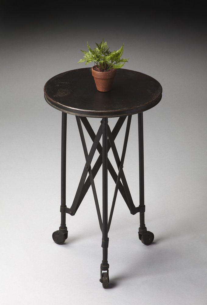 Round dark wooden-top end table on caster wheels with crisscross metal legs and a small potted fern.