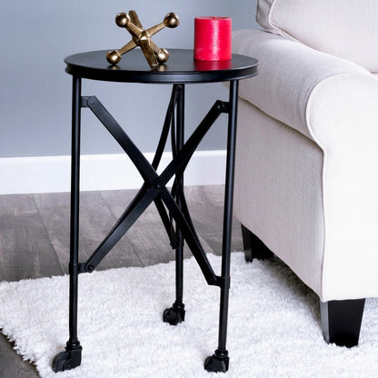Round black metal side table with casters and cross brace legs beside a beige sofa; red candle and brass sculpture on top.