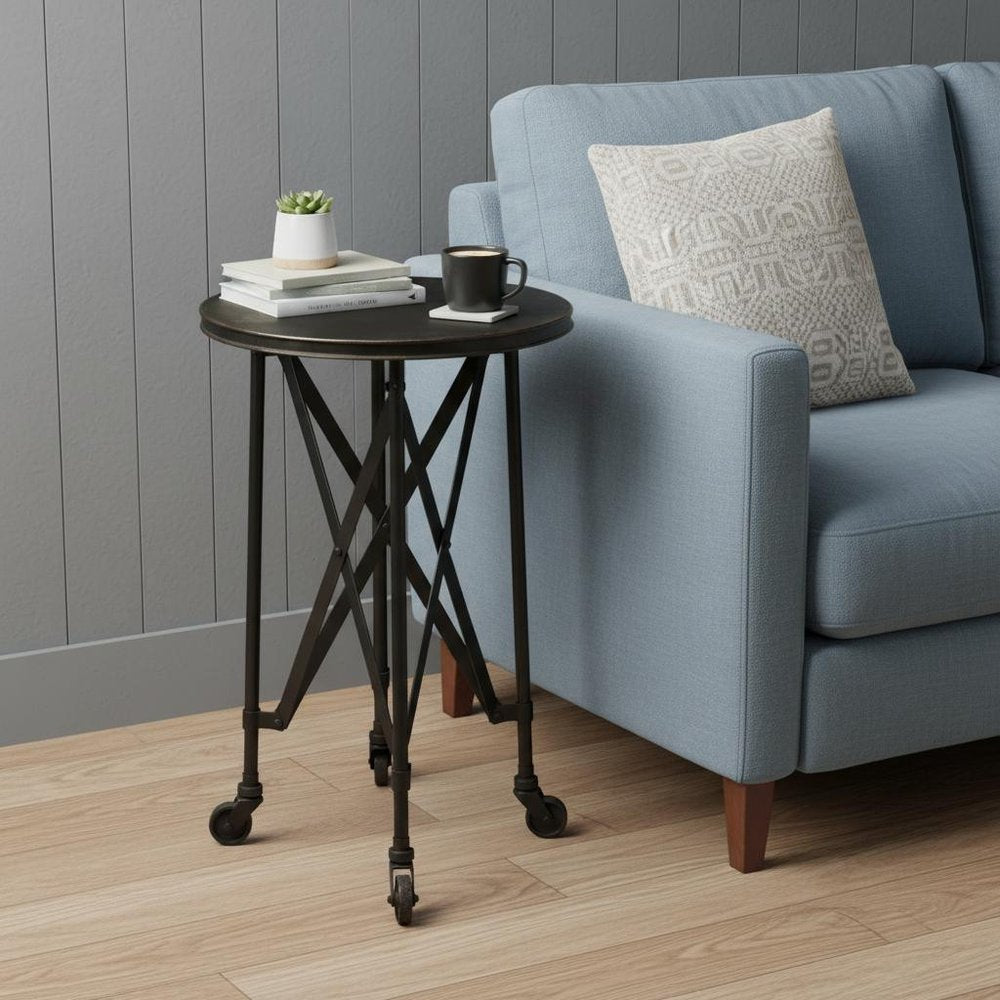 Round black metal rolling end table with crisscross legs beside a light blue sofa; books, plant, and mug on top.