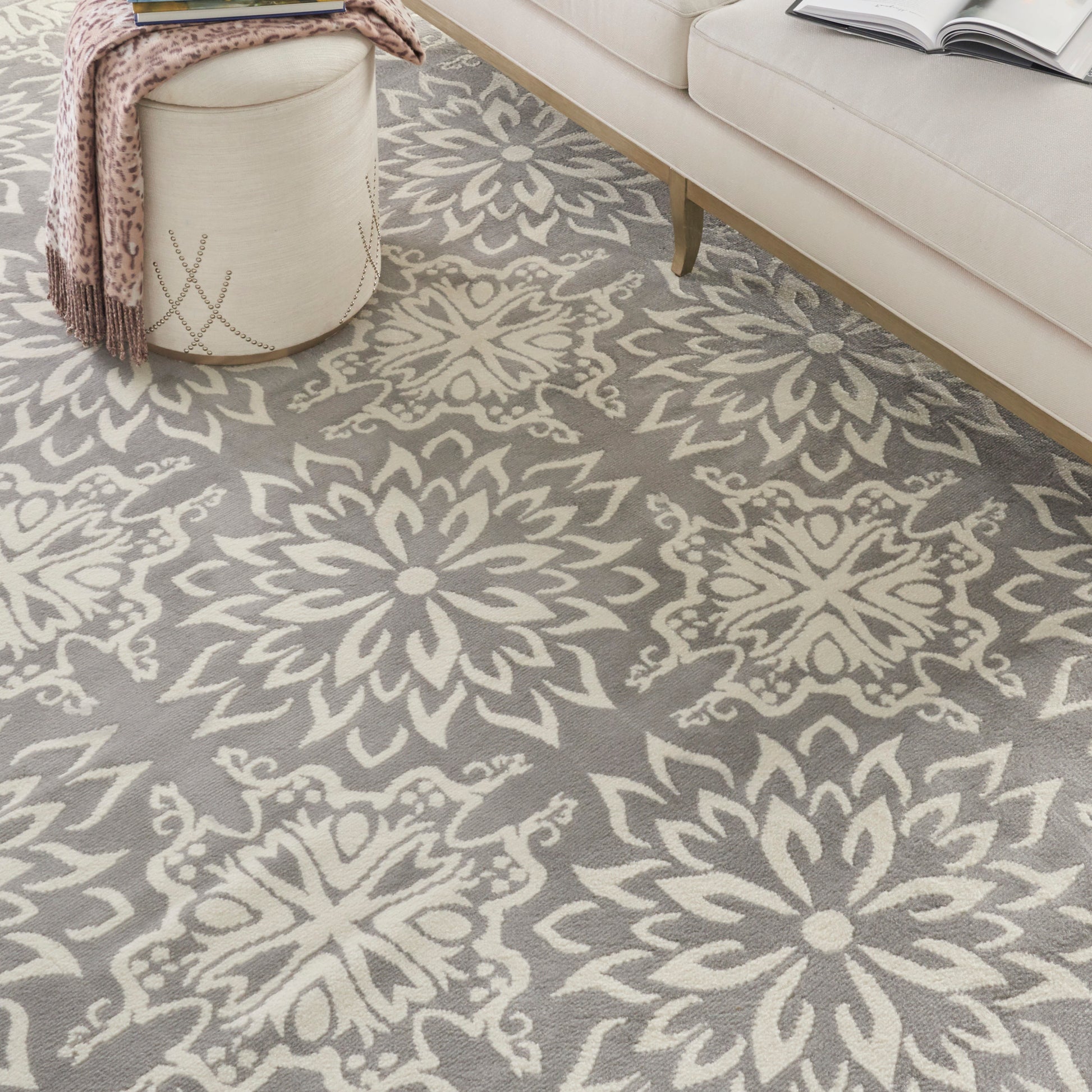 Grey rug with large cream floral damask pattern, cream sofa, and a knitted throw draped over a round ottoman.