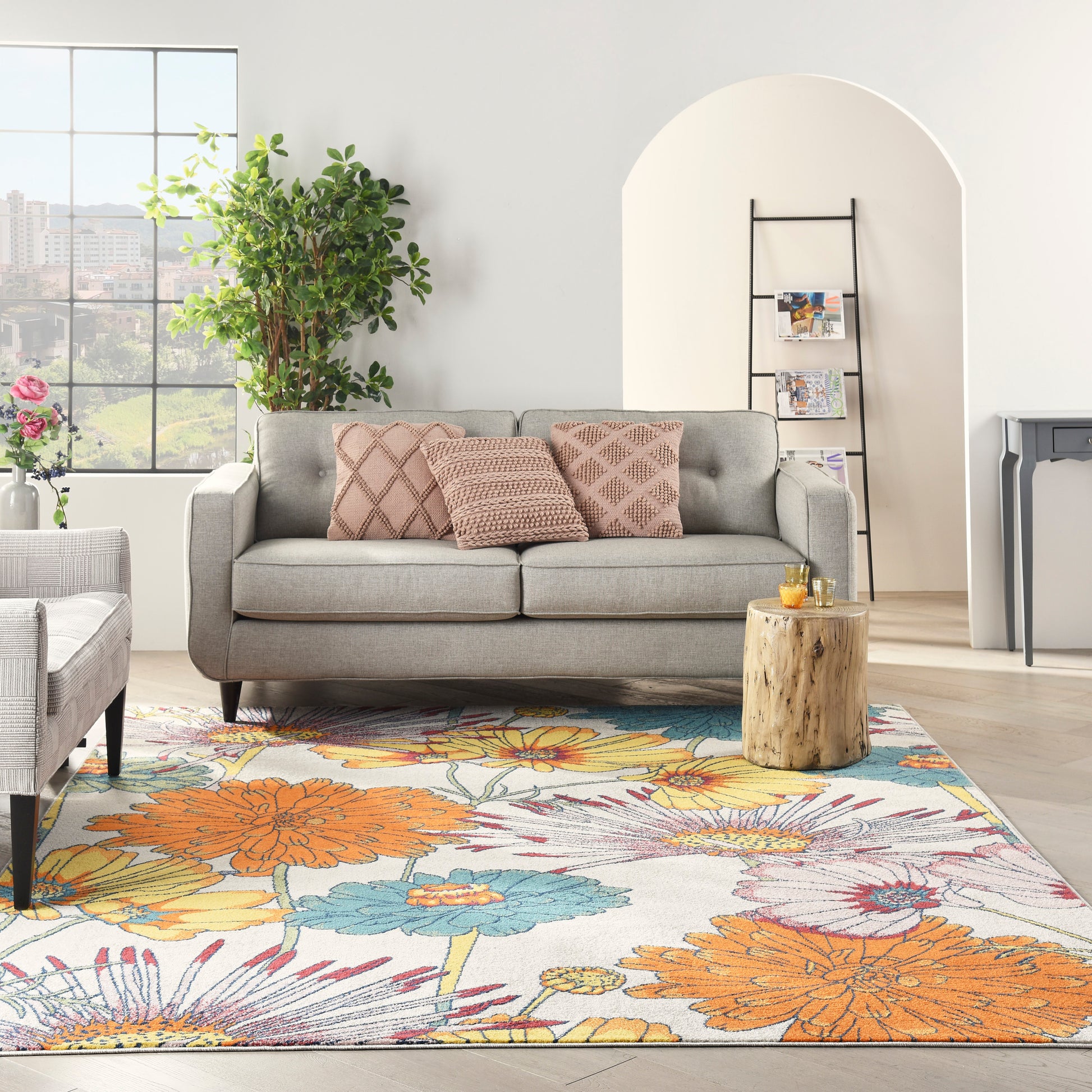 Cozy living room with gray sofa, pink pillows, vibrant floral rug, potted plant, and a wood stump side table.