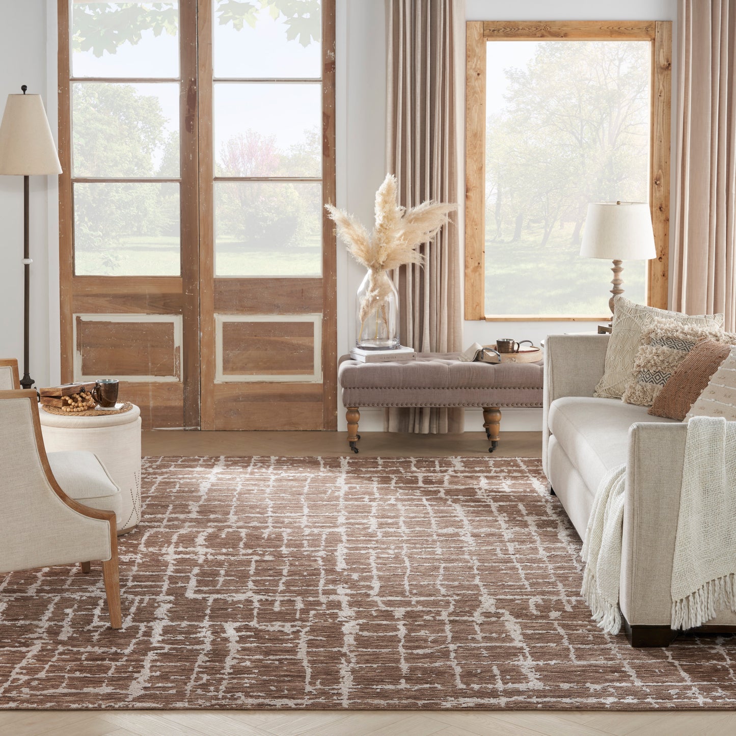 Cozy living room with beige sofa and pillows, wood-framed doors and large window, brown patterned rug, and pampas grass vase on an ottoman.