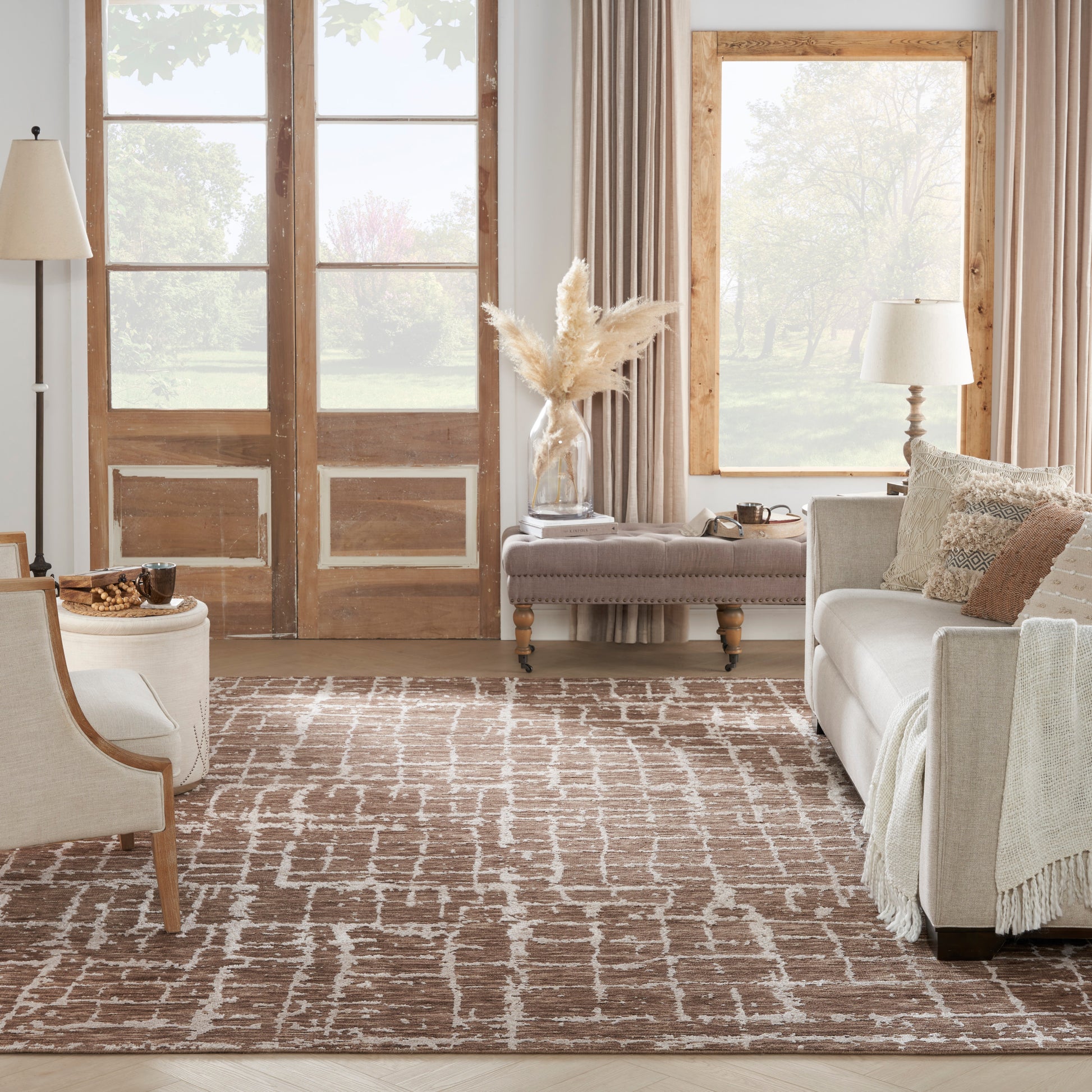Cozy living room with beige sofa and pillows, wood-framed doors and large window, brown patterned rug, and pampas grass vase on an ottoman.
