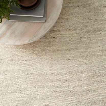 Beige textured woven rug with a round light wood coffee table and stacked books topped by a plant.