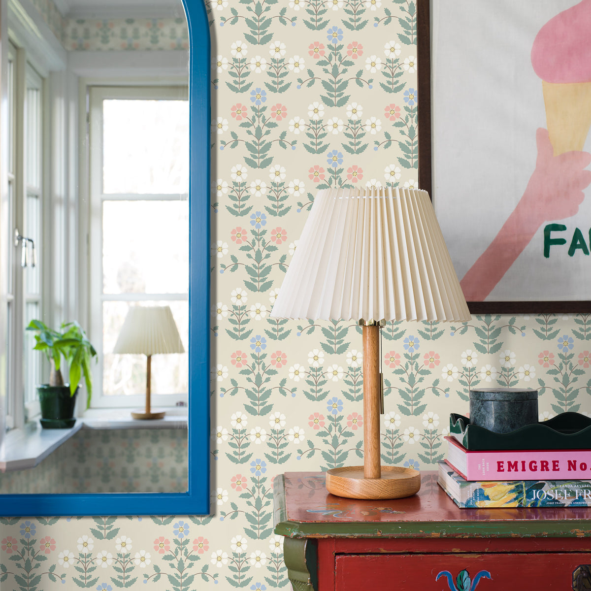 Vintage wooden table lamp with a pleated cream shade on a red console, beside stacked books against floral wallpaper.
