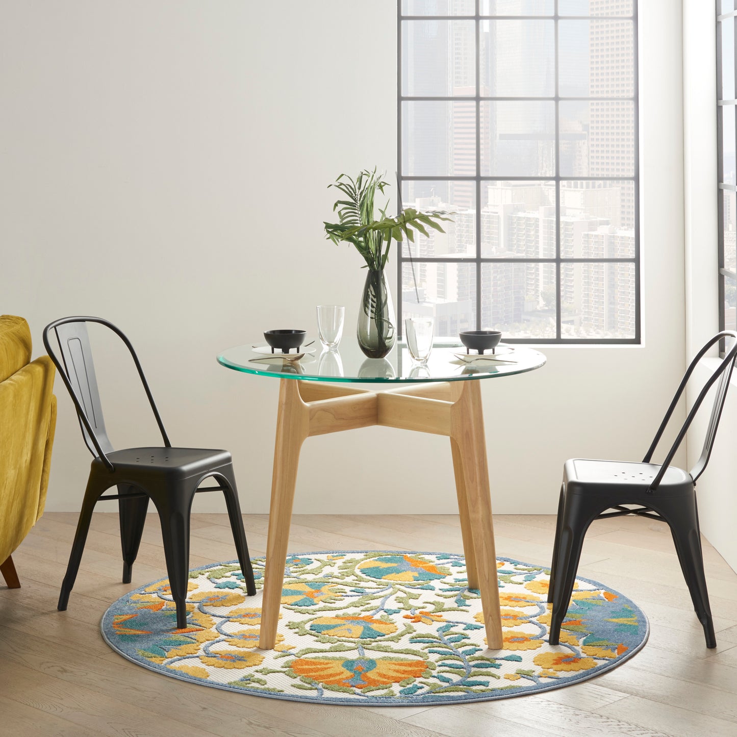 Round glass dining table with wooden base, two black metal chairs, colorful floral rug, vase of greenery, city skyline view.
