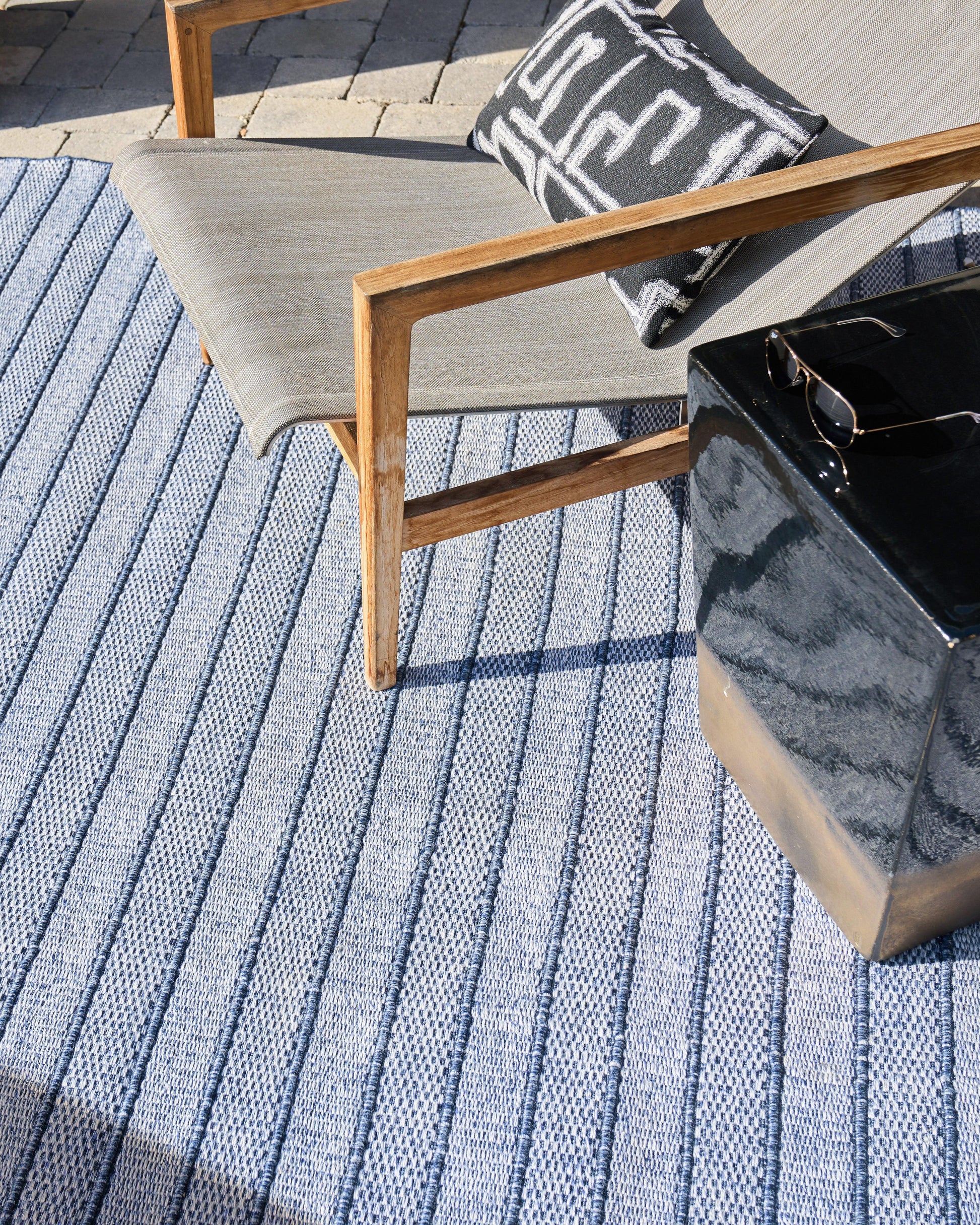 Flat-weave outdoor rug with blue stripes; visible in a setting with a gray-cushioned wooden lounge chair, a black-and-white pillow, and a glossy black side table.