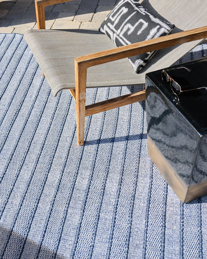 Flat-weave outdoor rug with blue stripes; visible in a setting with a gray-cushioned wooden lounge chair, a black-and-white pillow, and a glossy black side table.