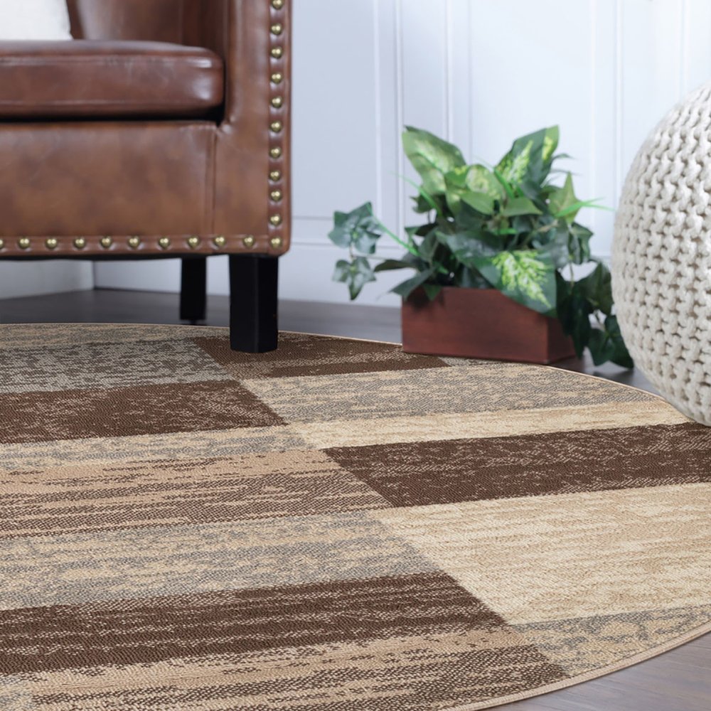 Round patchwork rug in beige, slate gray and brown with geometric blocks, placed under a brown leather sofa with studded trim, with a plant and woven pouf nearby.