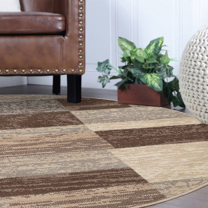 Round patchwork rug in beige, slate gray and brown with geometric blocks, placed under a brown leather sofa with studded trim, with a plant and woven pouf nearby.
