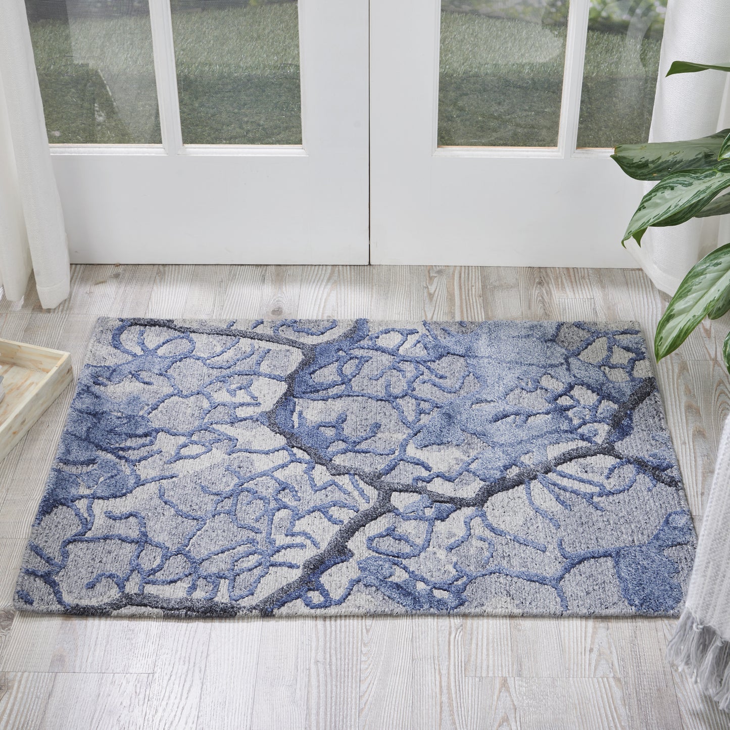 Blue-gray abstract rug with tree-branch pattern on light wood floor by glass doors; plant visible at edge.