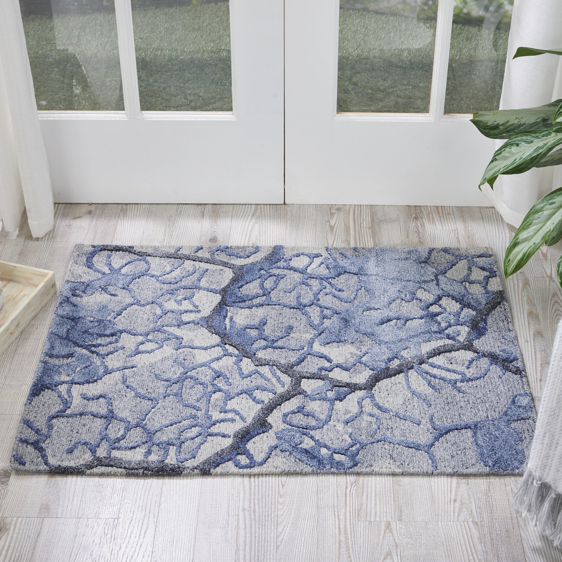 Blue-gray abstract rug with tree-branch pattern on light wood floor by glass doors; plant visible at edge.